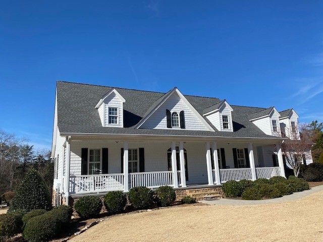 White house with a wraparound porch, black shutters, and dormers, under a blue sky.