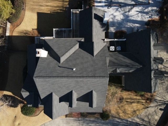 Overhead view of a house with a dark gray shingle roof and multiple gables, surrounded by trees and a brown lawn.