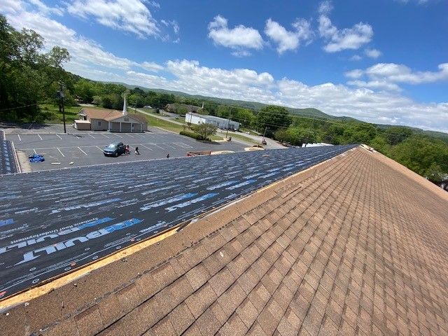 Roof partially covered with asphalt shingles; a section shows underlayment. Bright sunny day, view of a parking lot and hills.