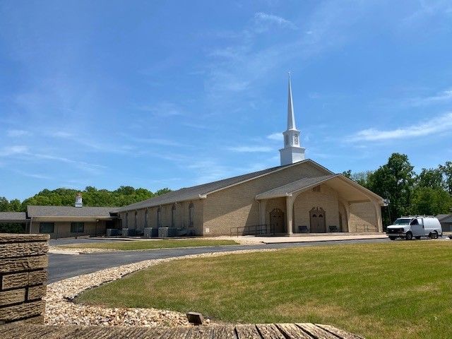 A light-colored brick church with a white steeple under a blue sky, a van parked nearby.