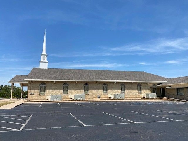 Church building with a white steeple, beige brick exterior, and a parking lot on a clear, sunny day.