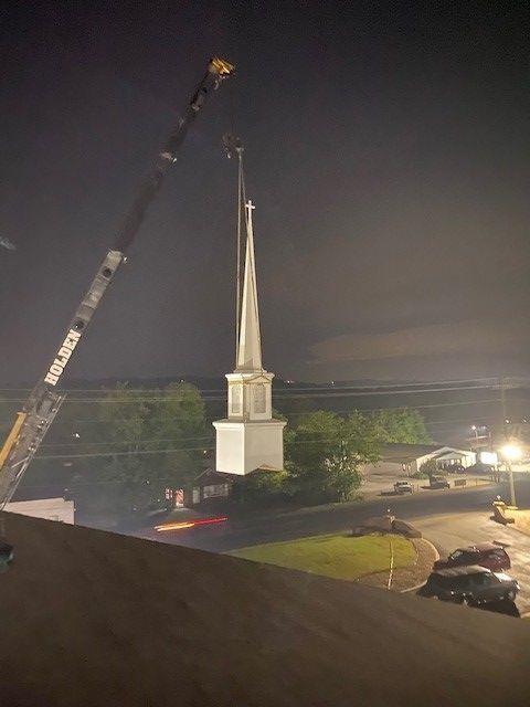 A crane lifting a church steeple at night. A dark sky surrounds the white building.