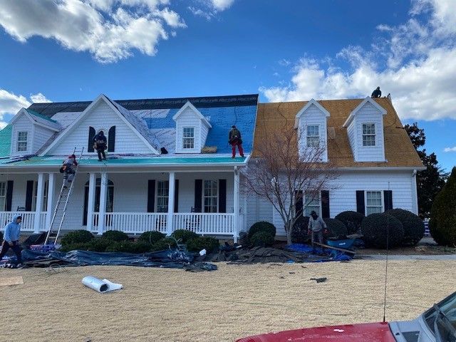 Roofers working on a white house, covering the roof with blue and brown materials, under a blue sky.