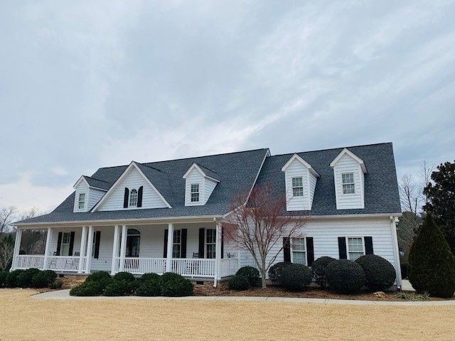 White house with a gray roof, black shutters, and three dormer windows under a cloudy sky.