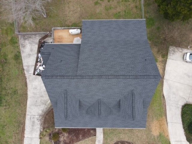 Overhead view of a dark gray shingled roof with a concrete driveway and small building in the background.