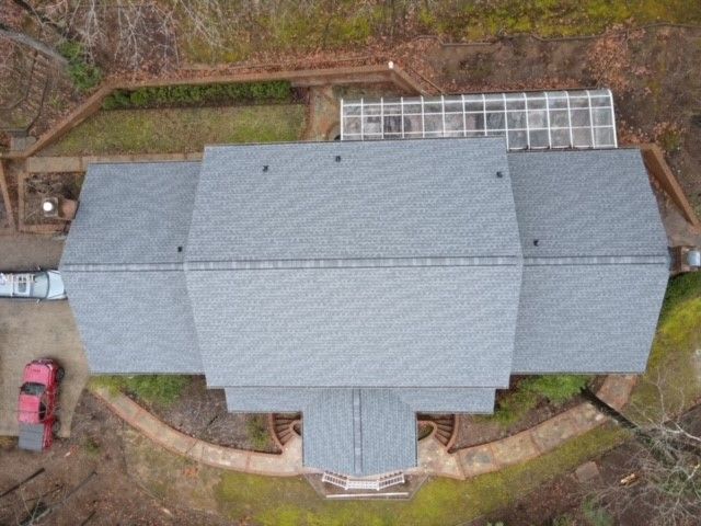 Overhead view of a gray-roofed house with a greenhouse, red car in driveway, surrounded by a curving pathway.