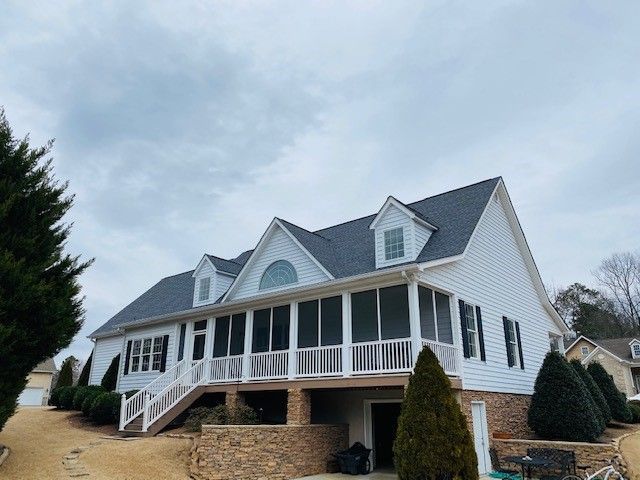 White house with a screened porch, stone foundation, and dark gray roof under an overcast sky.