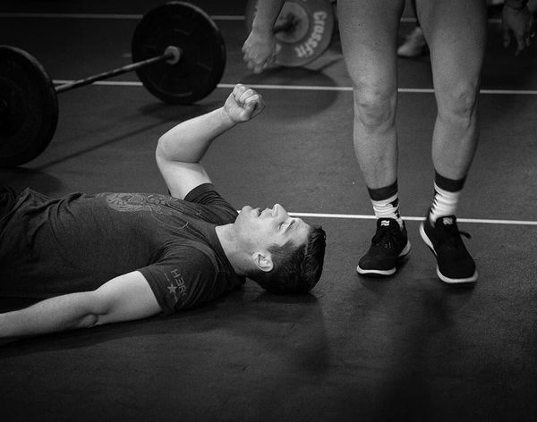 Person lying on gym floor beside a barbell, reaching a hand toward another person standing nearby.