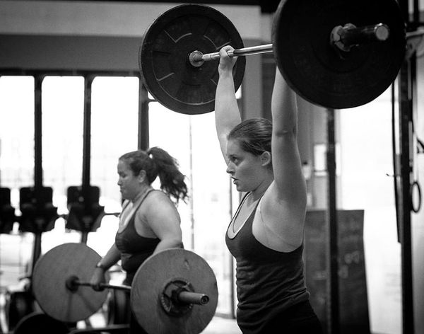 Two people lifting barbells overhead in a gym in Surrey, BC, one in the foreground and one behind.