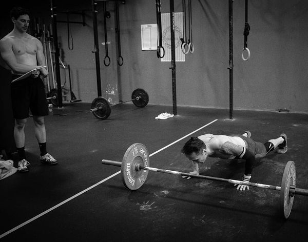 Athlete doing a push-up beside a barbell in a gym in Surrey, BC, with another person standing nearby.