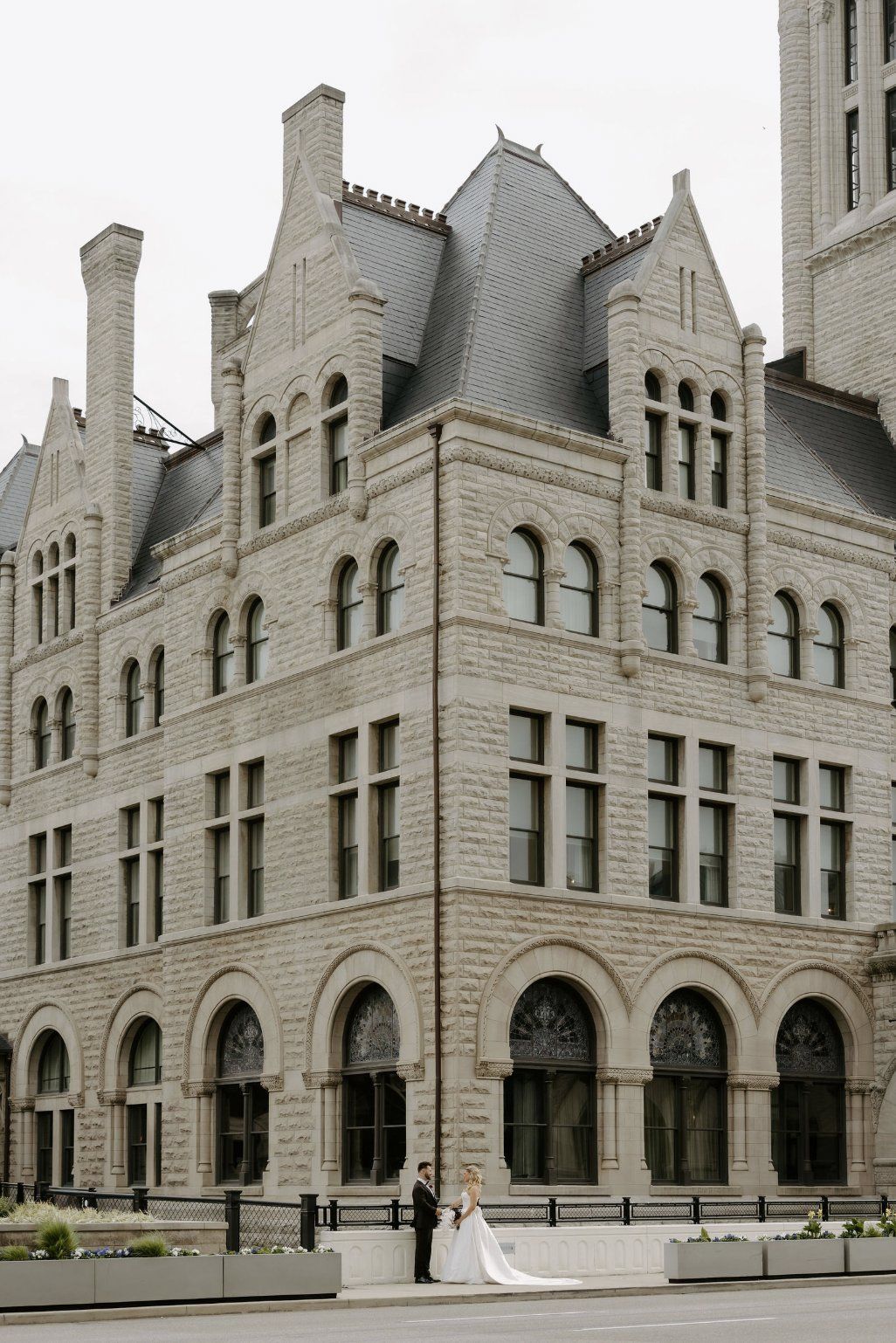 Newlyweds pose in front of a stone building with arched windows, slate roof by Luxury Wedding Planners in Nashville, TN