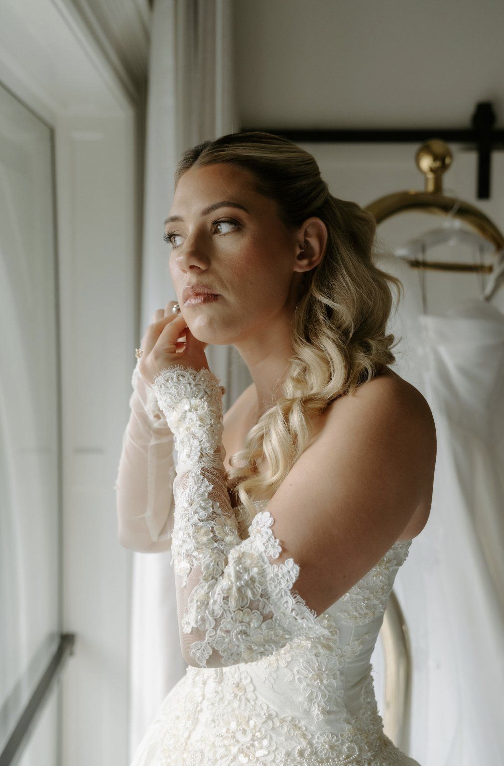 Bride in a detailed white wedding gown looks out a window, adjusting an earring by Luxury Wedding Planners in Nashville, TN