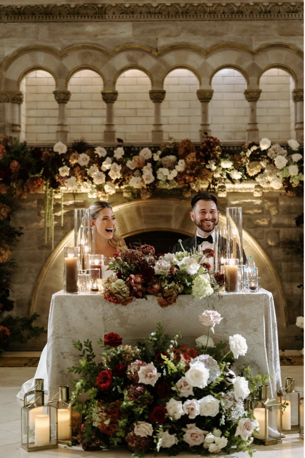 Couple at a wedding reception, seated at a decorated table, smiling. Stone archway backdrop with floral arrangements by Luxury Wedding Planners in Nashville, TN