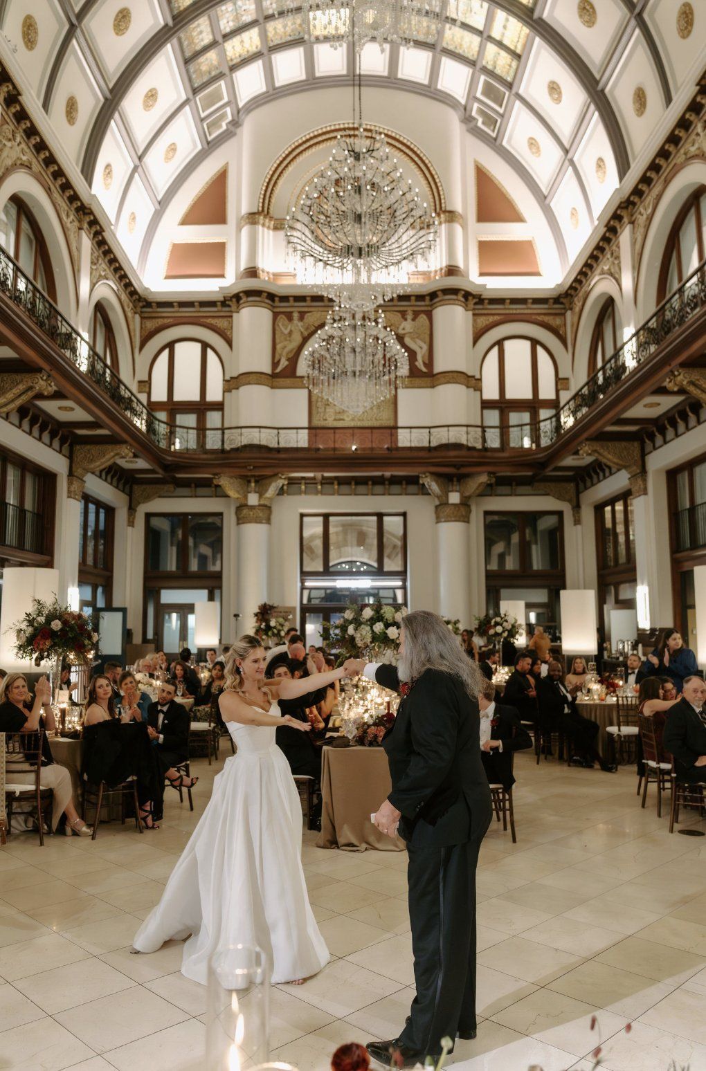 Bride dancing with a man in an ornate ballroom. High ceilings, large chandelier, wedding guests by Luxury Wedding Planners in Nashville, TN
