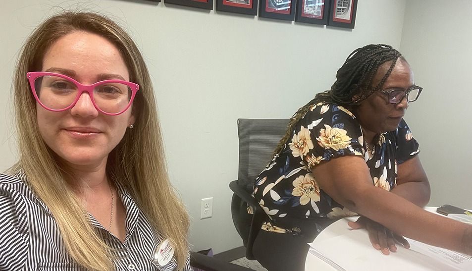 Woman with pink glasses smiles at the camera; another woman works at a desk in an office.