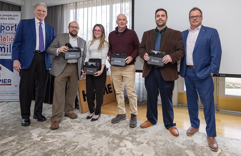 Group of six people holding devices, posing for a photo indoors. Some wear suits, smiling.