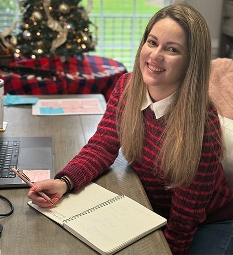 Woman smiles, writing in a notebook at a desk. Red striped sweater, white collared shirt. Christmas tree in the background.