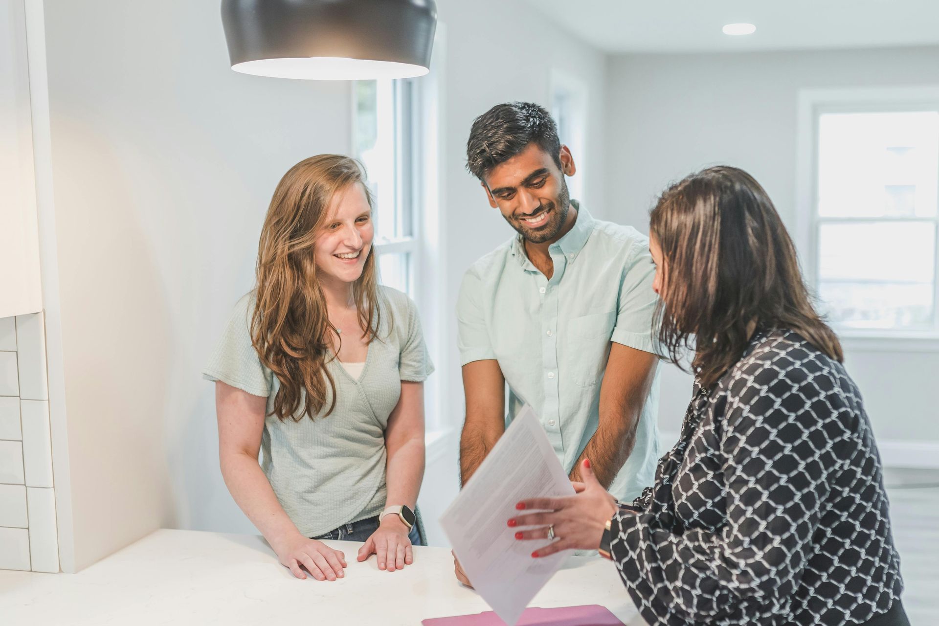 A realtor showing a document to a smiling couple in a bright, modern kitchen.