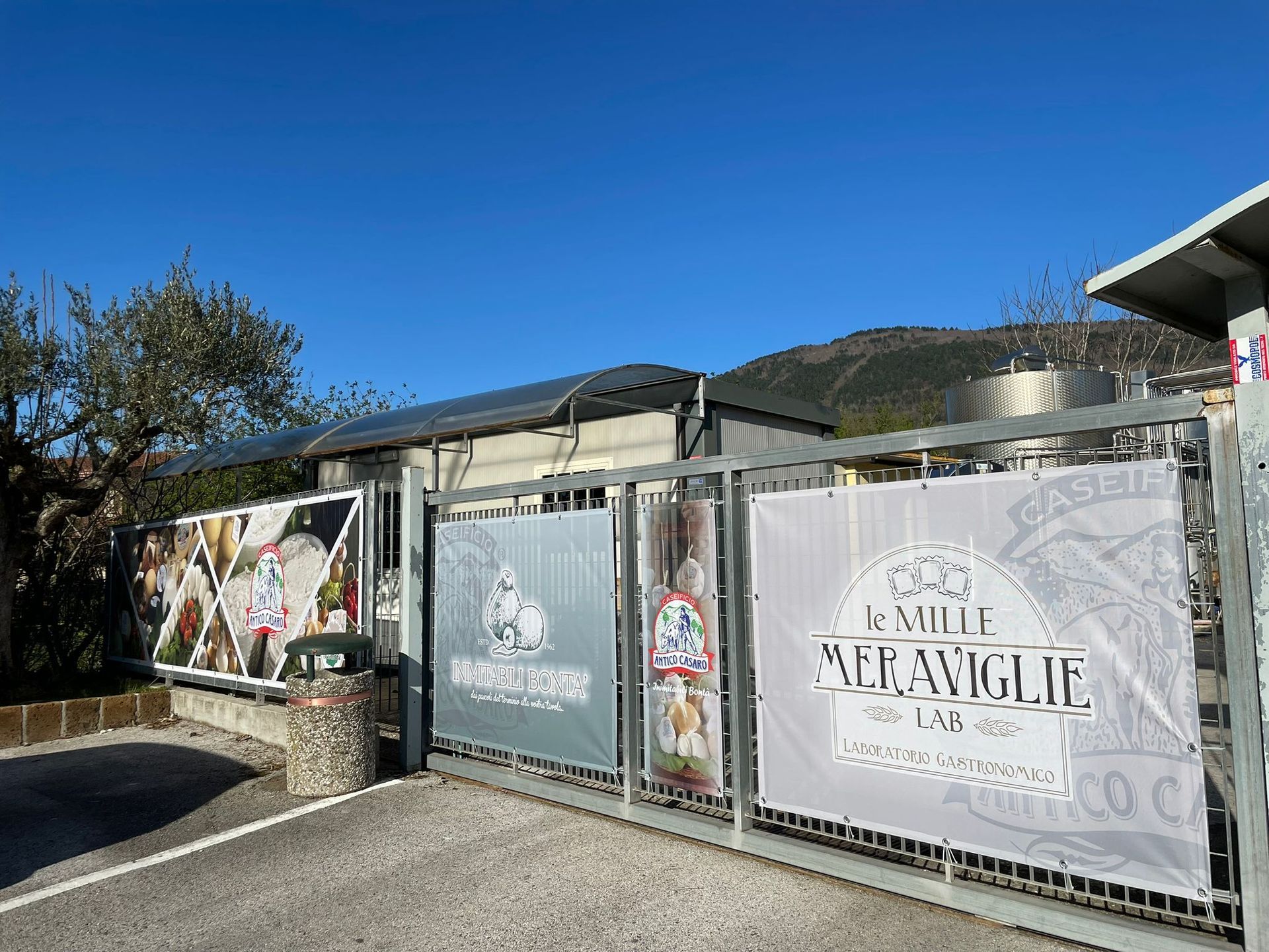 Cancello d'ingresso con striscioni per "Le Mille Meraviglie" di fronte a un edificio sotto un cielo azzurro.