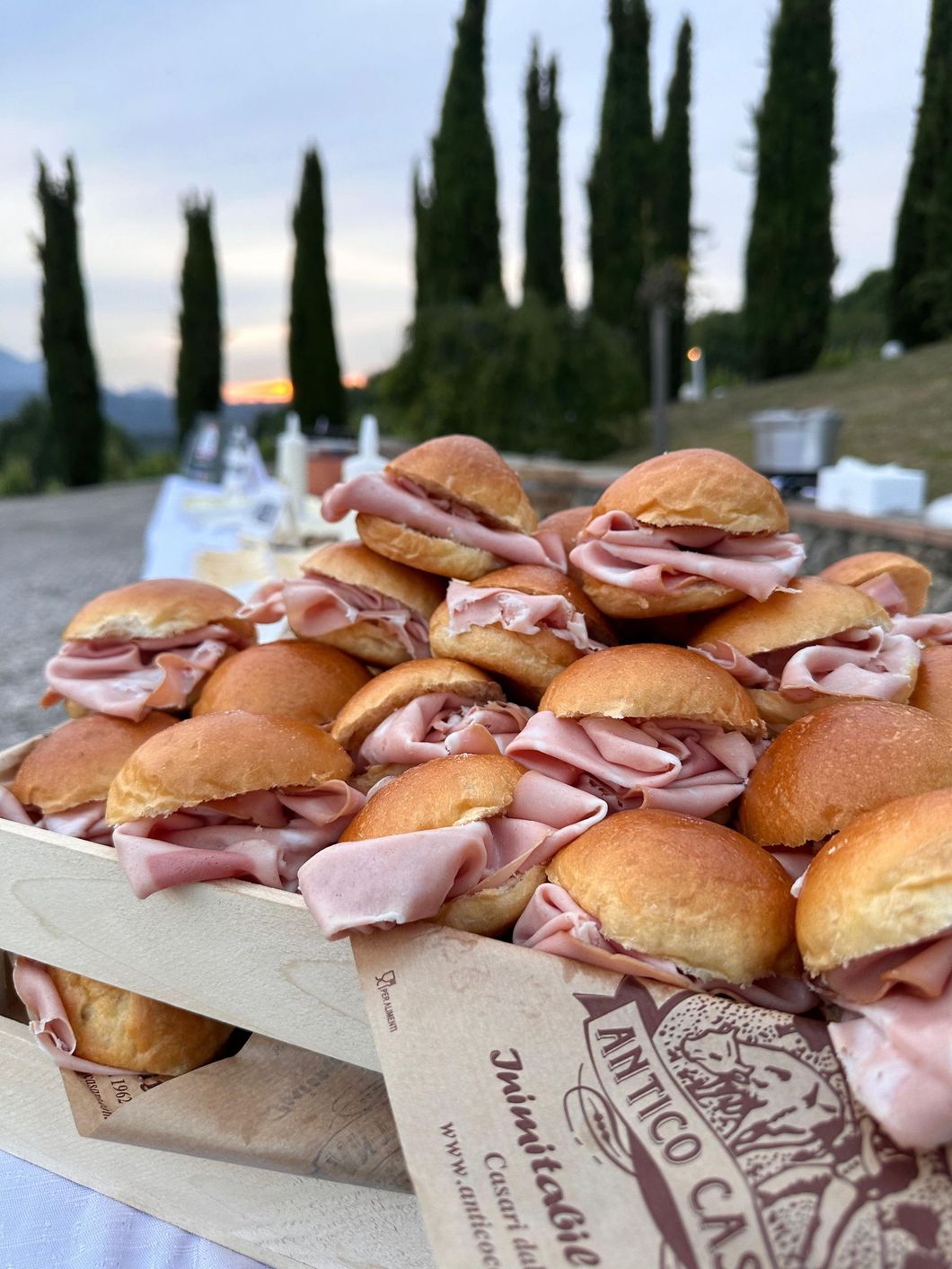 Mini hamburger di prosciutto su un vassoio di legno, disposti all'aperto con cipressi sullo sfondo.