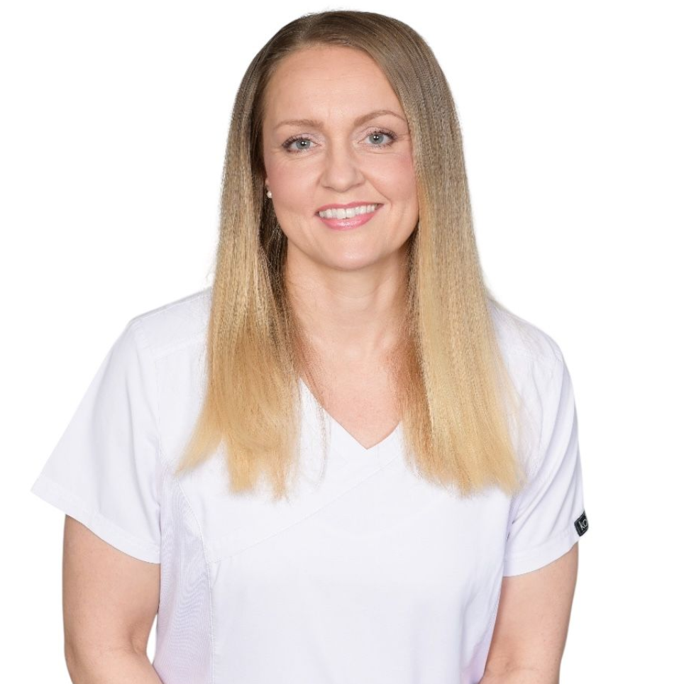 Kvinna med långt blont hår ler, klädd i en vit v-ringad scrub-topp.