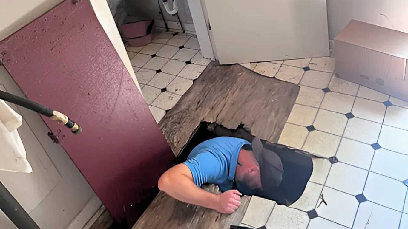 Man in blue shirt and hat reaching into hole in floor of room with checkered tiles and debris. Man in blue shirt and hat reaching into hole in floor of room with checkered tiles and debris.