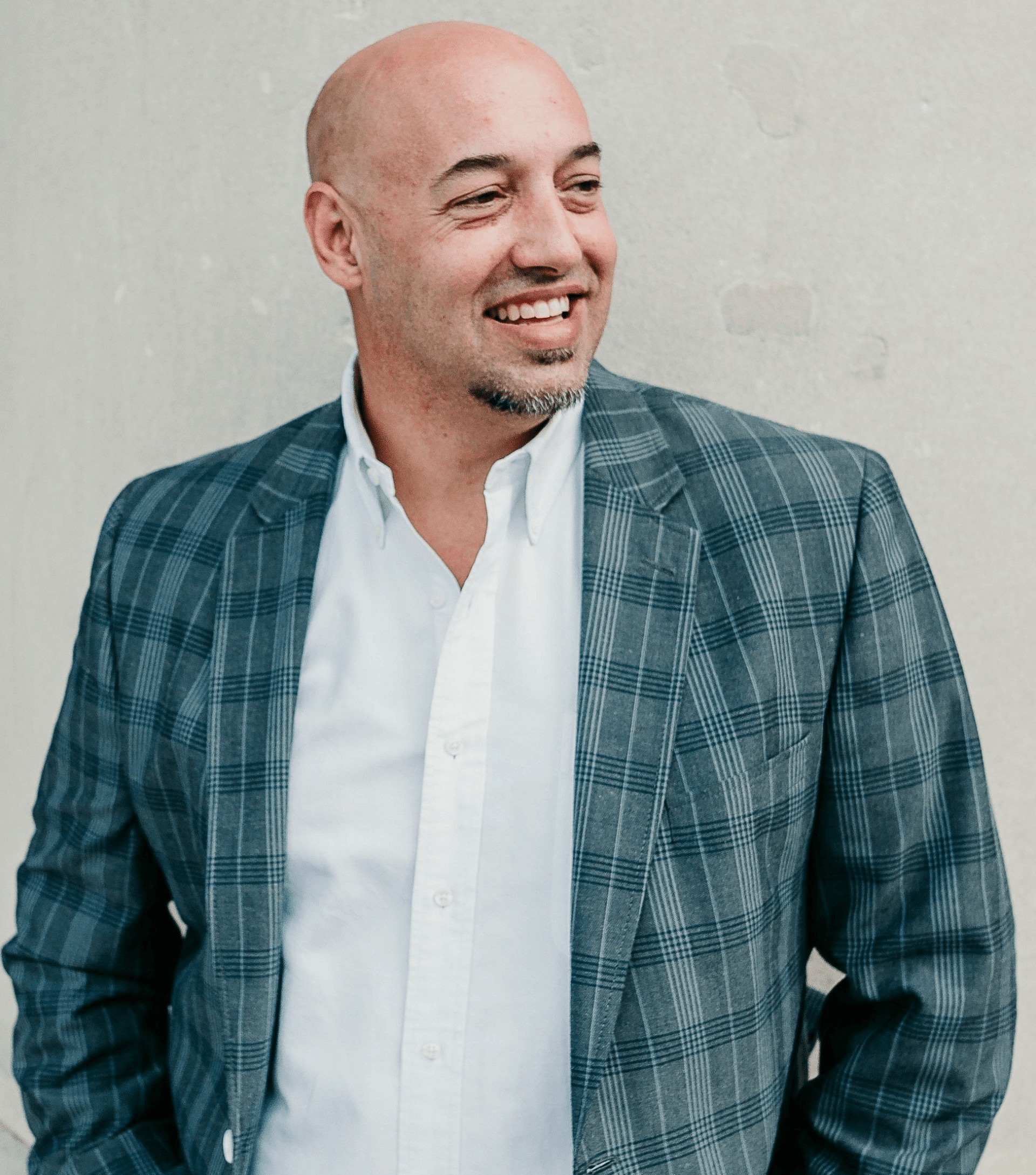 Smiling bald man in plaid blazer and white shirt, leaning against a wall.