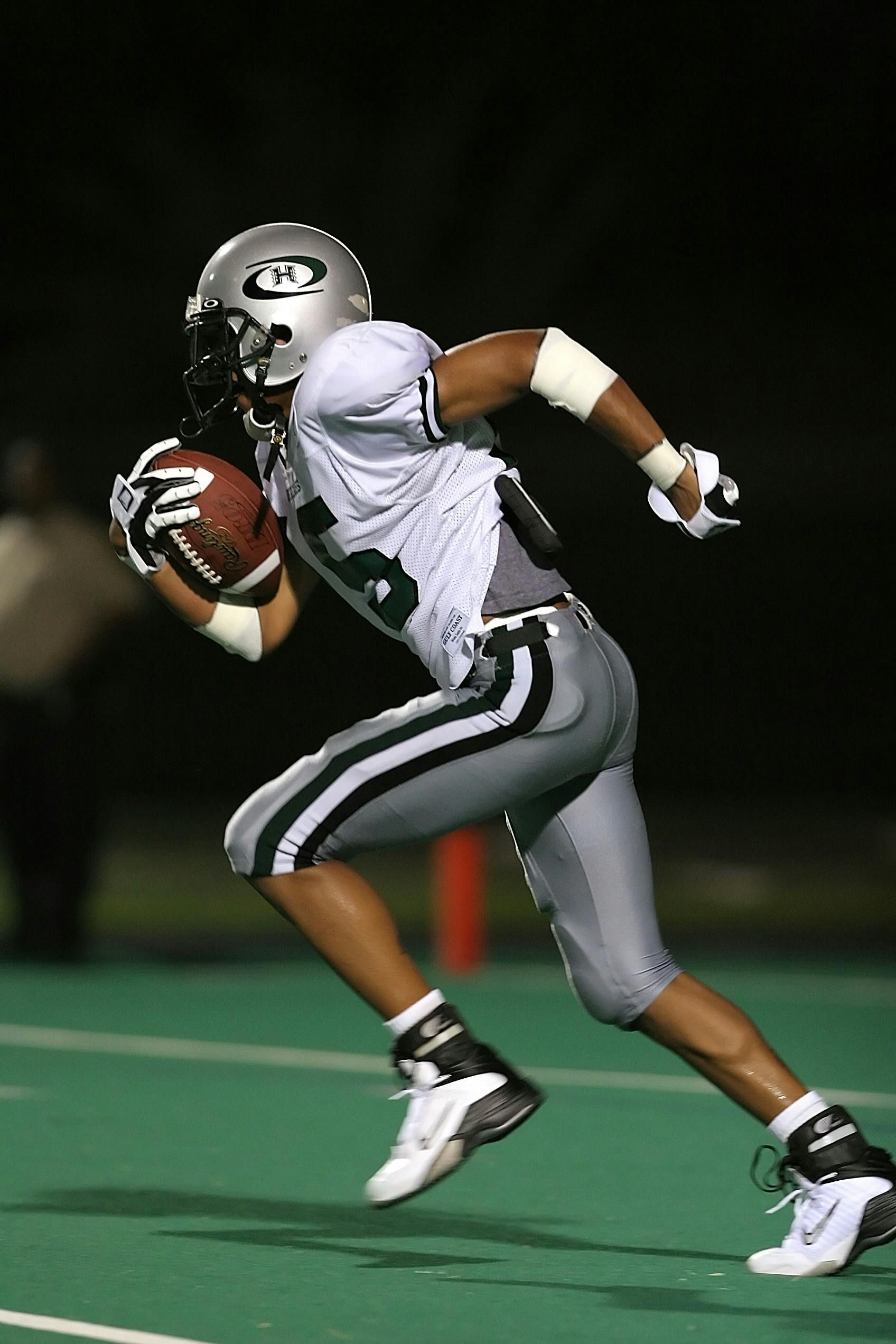 Football player in gray uniform running with the ball on a green field at night.
