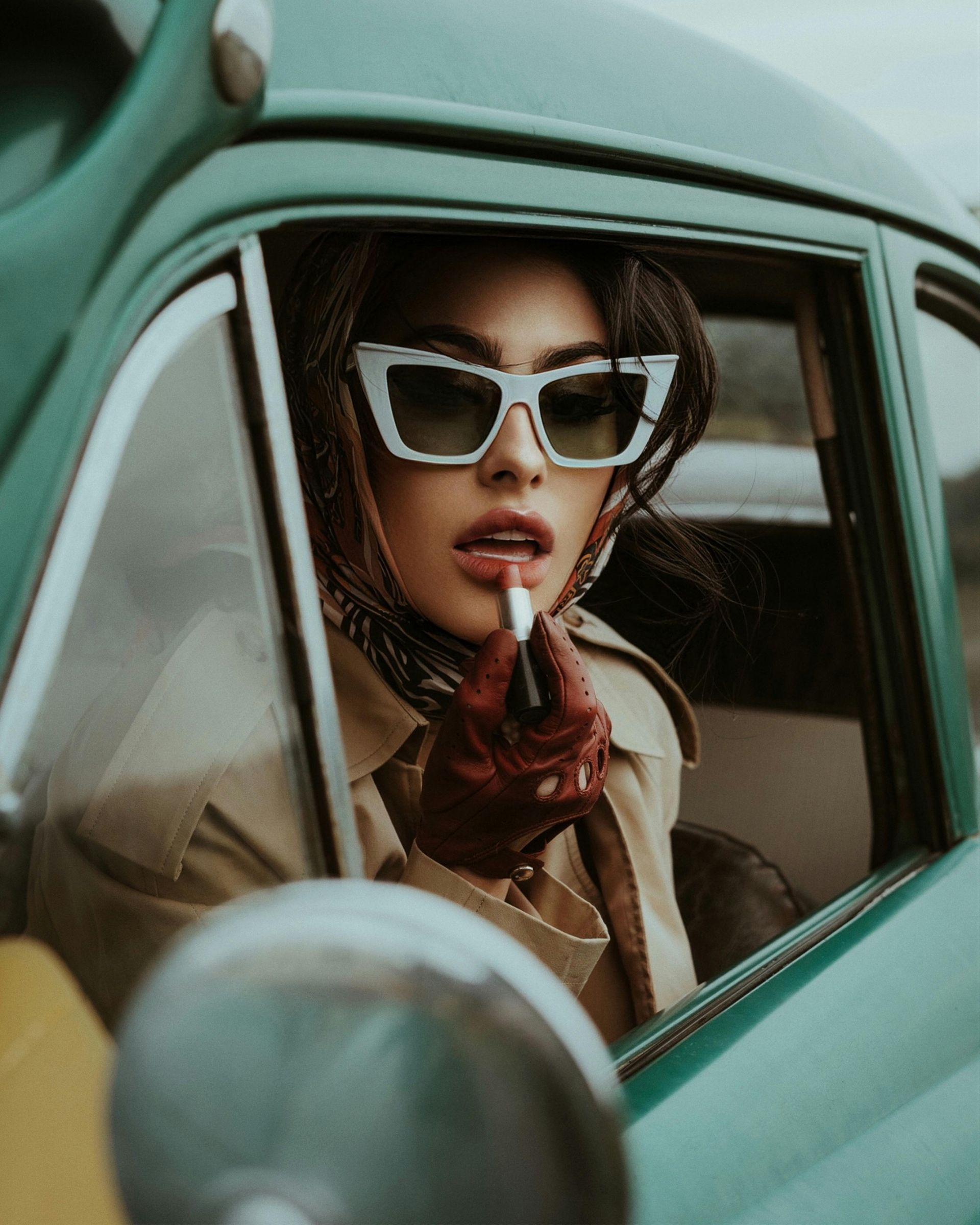 A woman wearing sunglasses is applying lipstick in a car.