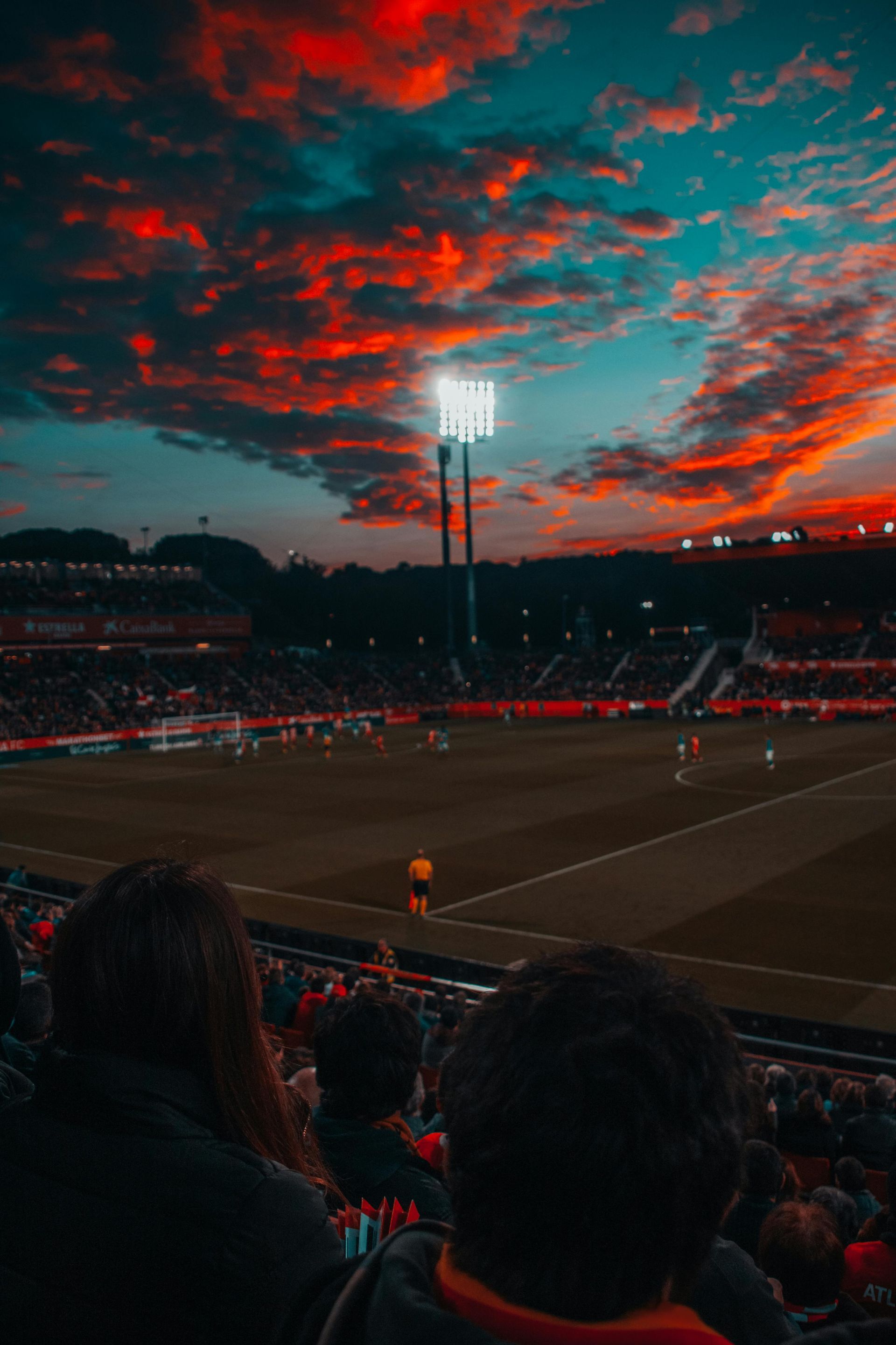 Soccer game under a fiery sunset, with a packed stadium and floodlights.