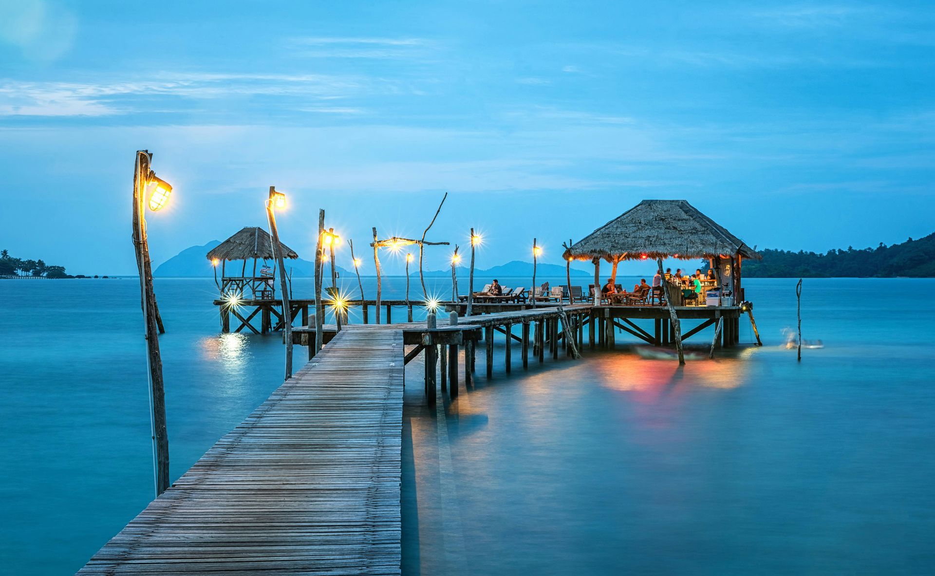 Wooden pier with thatched-roof huts extending over calm blue water at dusk, lit by warm lights.