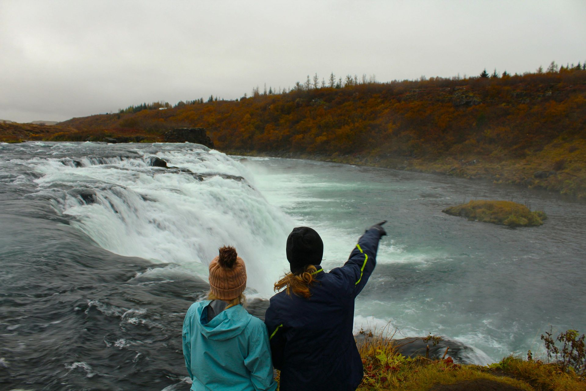 Two people at a waterfall; one points to it. Overcast sky, fall foliage.