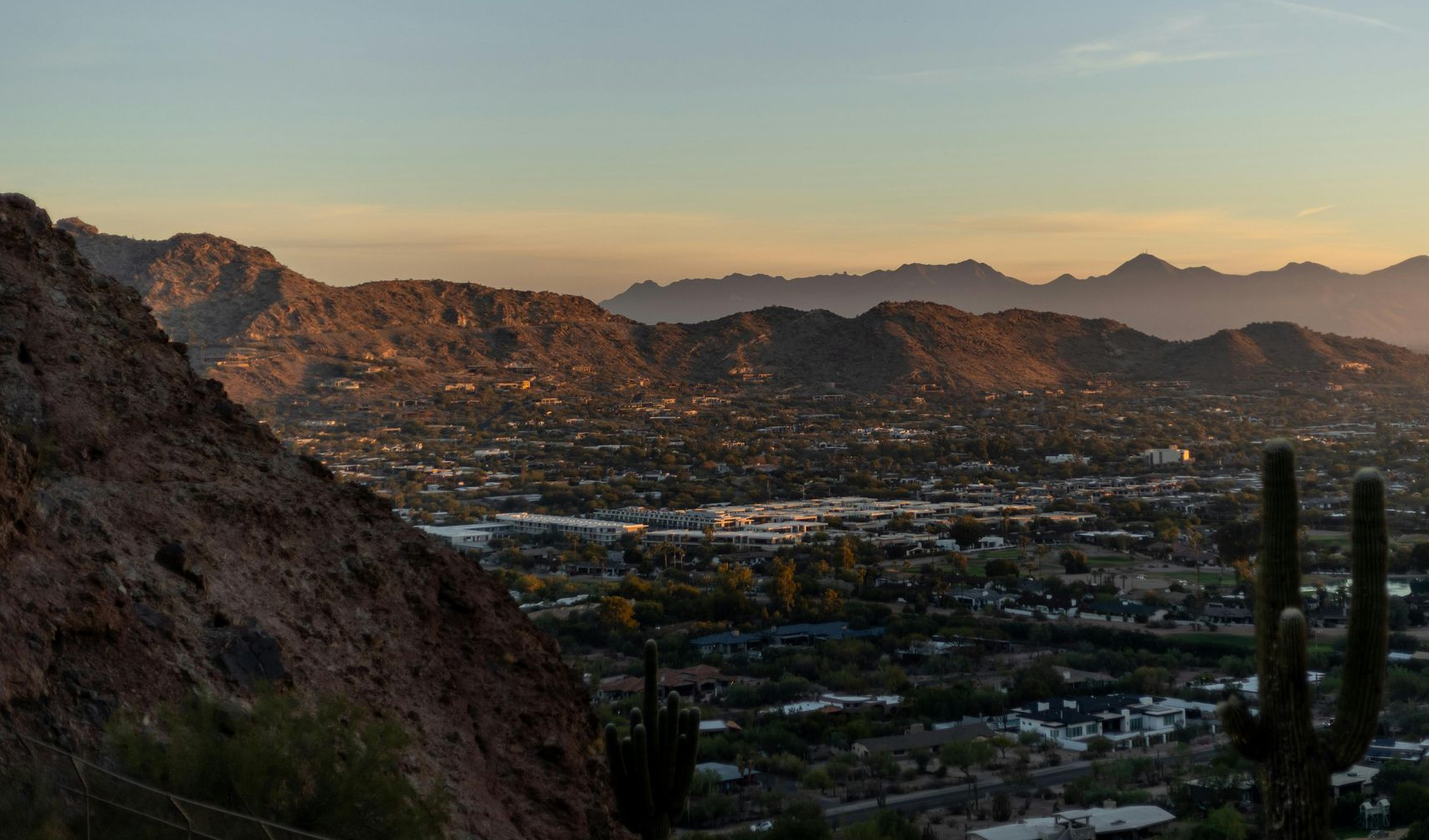 Valley view at dusk, illuminated city nestled between mountains, saguaro cactus in foreground, golden light.