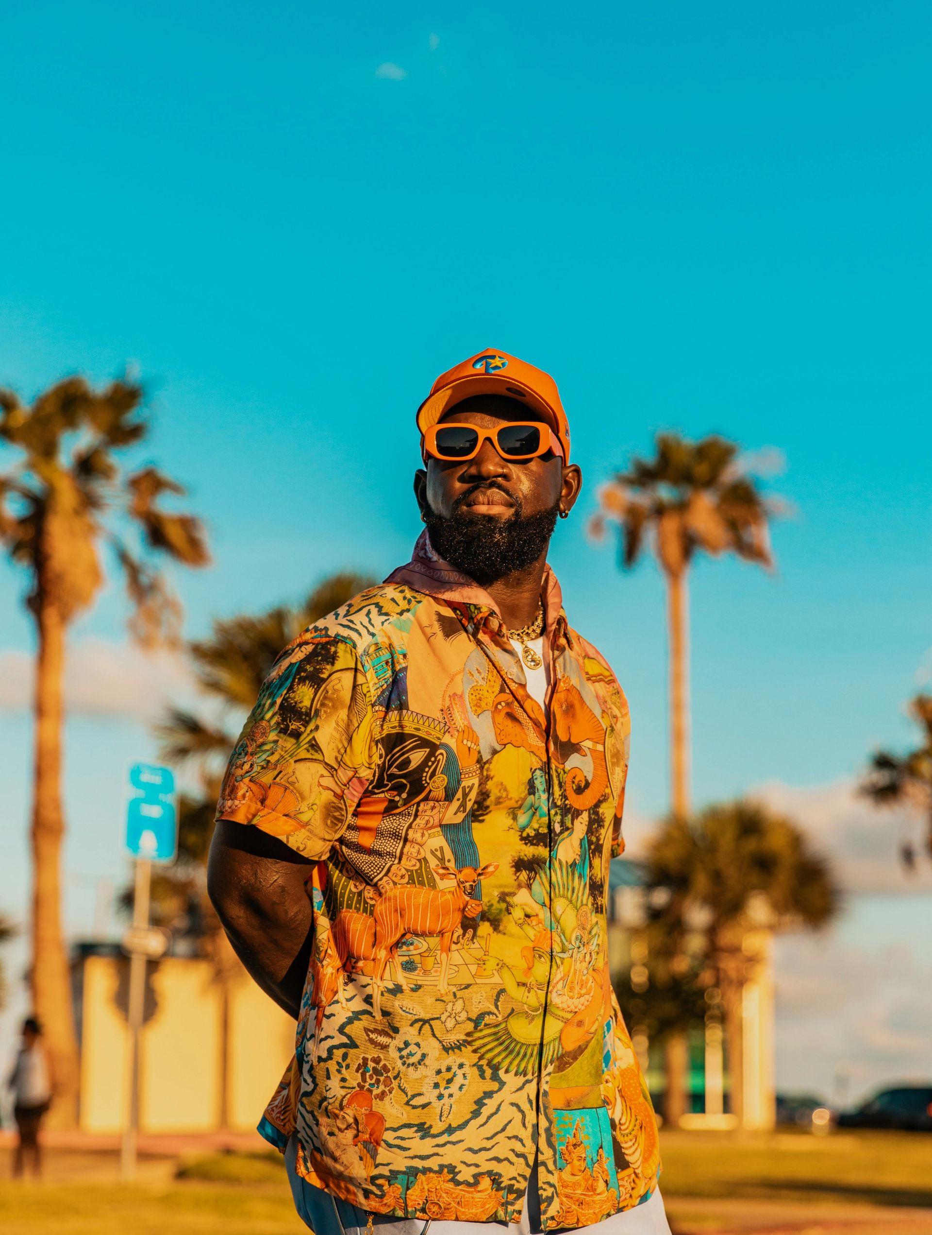 Man in a colorful shirt and sunglasses stands outdoors, palm trees and blue sky in the background.