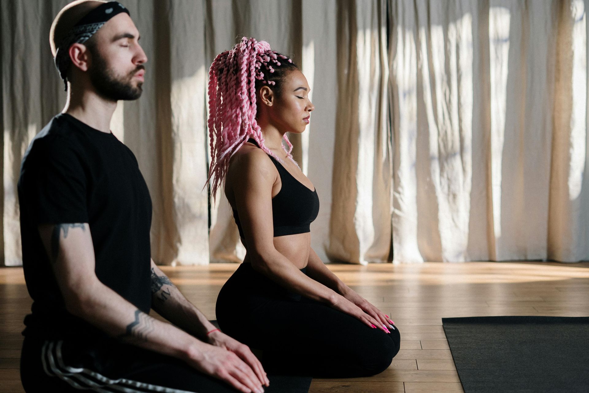 Two people meditating in a room with sunlight. Man wears black, woman has pink braids.