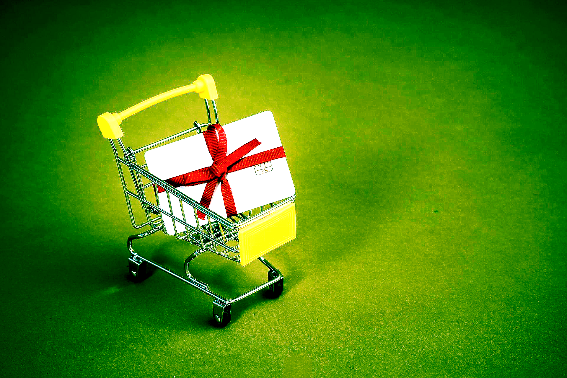 A small shopping cart holds a white gift box tied with a red ribbon on a green surface.