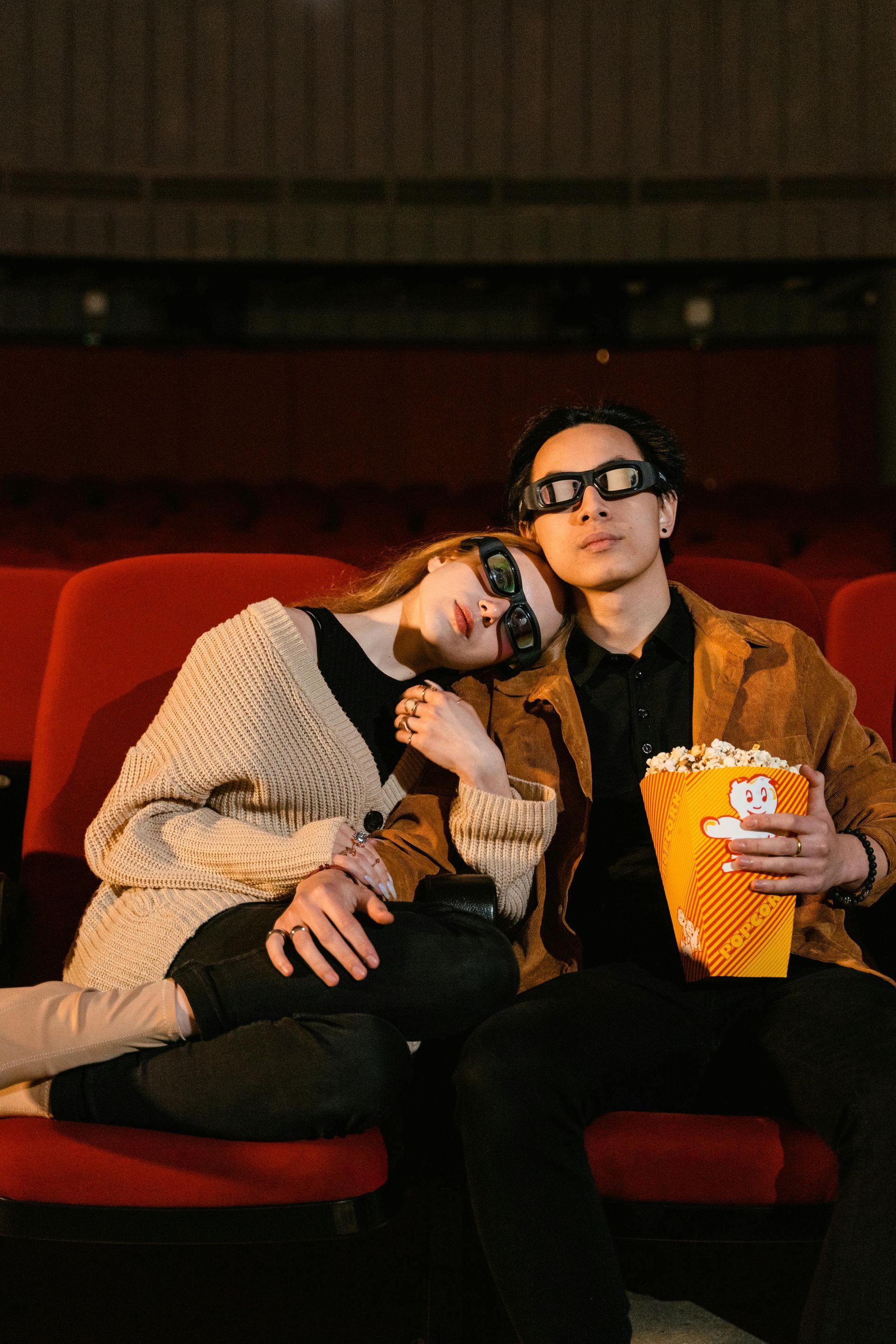 Couple in 3D glasses in a movie theater. Woman resting on man's shoulder, holding popcorn. Red seats, dark setting.