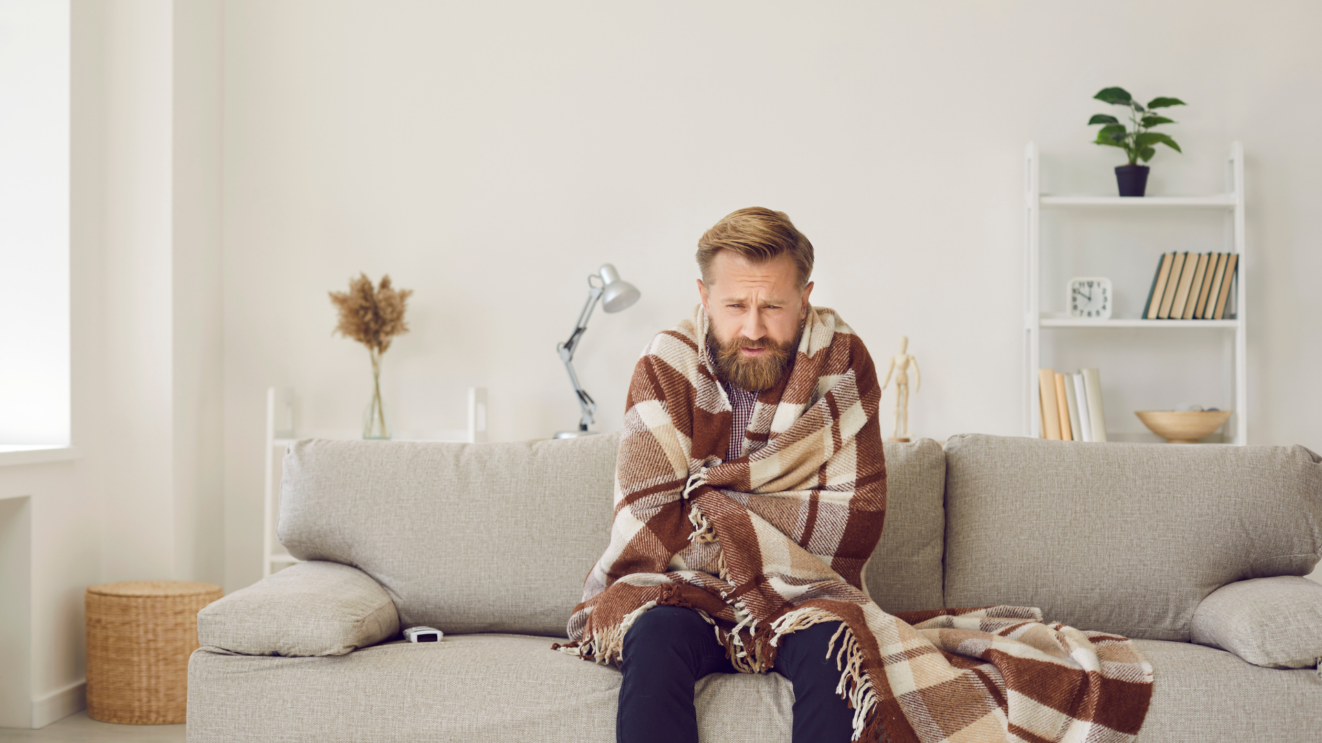 Man wrapped in blanket sits on a couch, looking cold. Living room setting.
