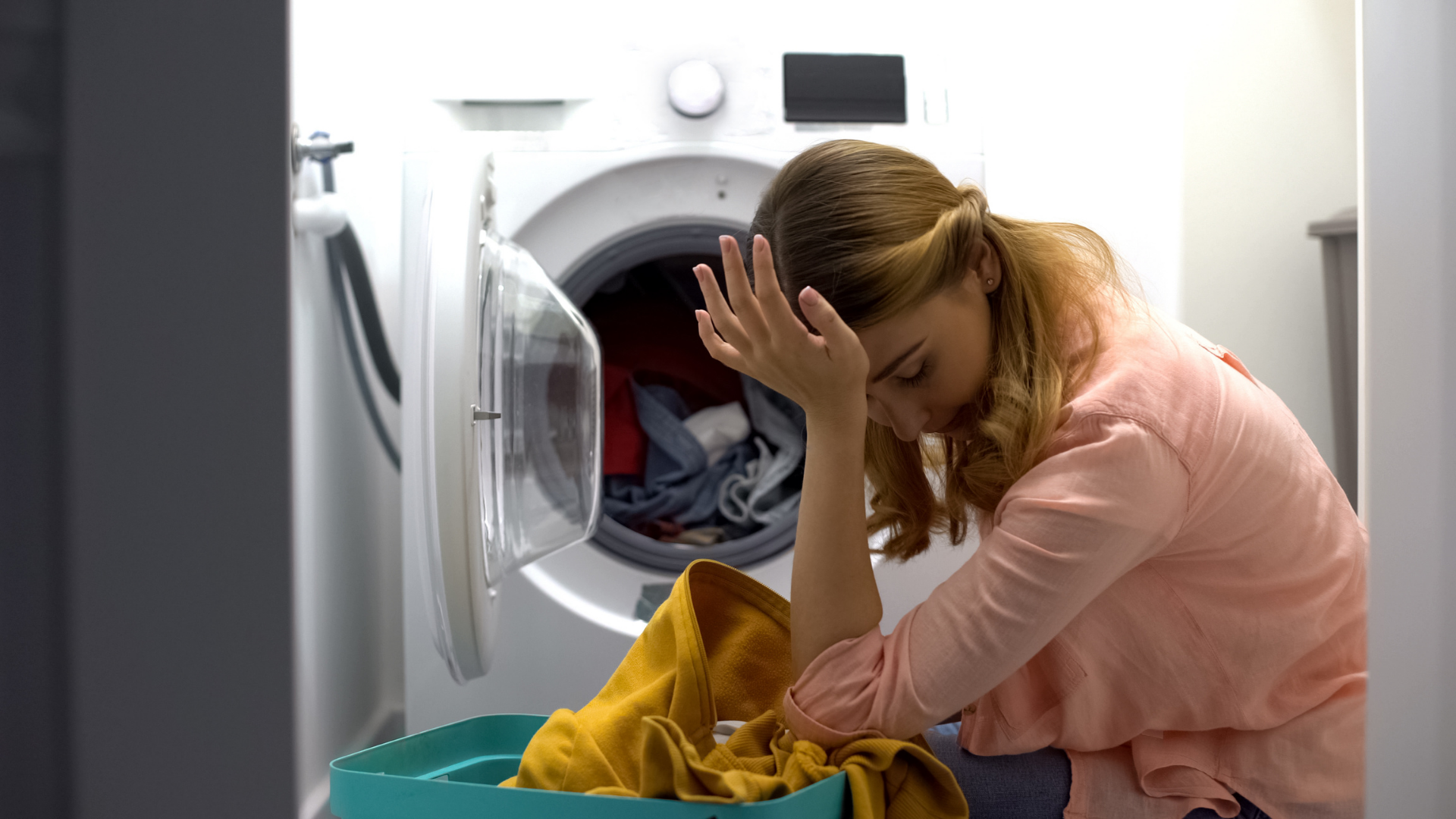 Woman with hand on forehead, looking dejected, sits next to washing machine and laundry basket with clothes.