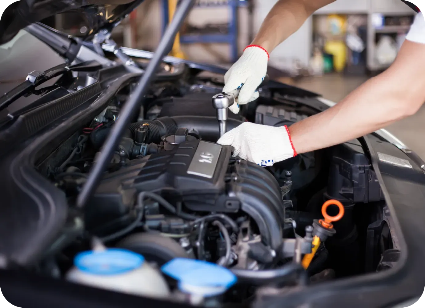 A mechanic wearing white gloves uses a ratchet tool to work on a car engine inside a garage.