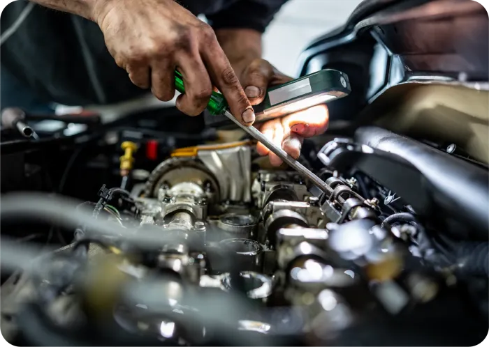 A mechanic uses a screwdriver and a portable light to work on the internal components of a car engine.