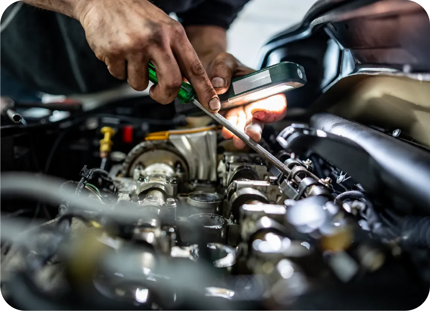 A mechanic uses a screwdriver and a handheld work light to perform maintenance on an open car engine.