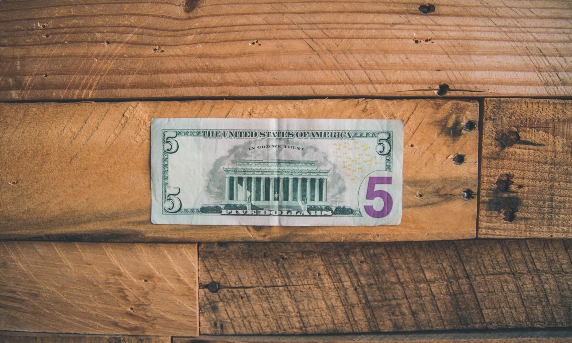 A five-dollar bill sits on a wooden surface.