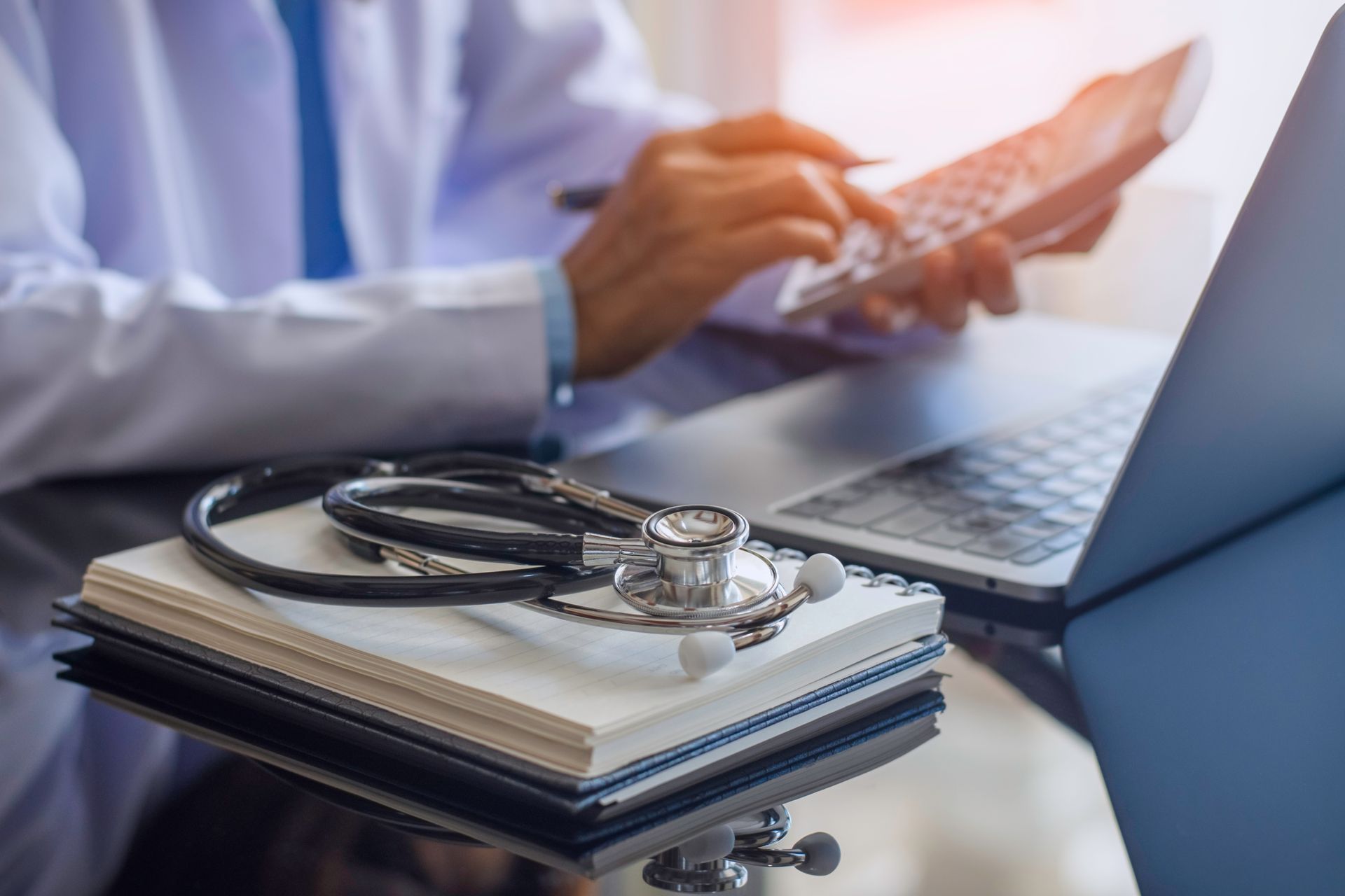 A partial view of a doctor using a calculator & a laptop, with a stethoscope & a notebook on a desk.