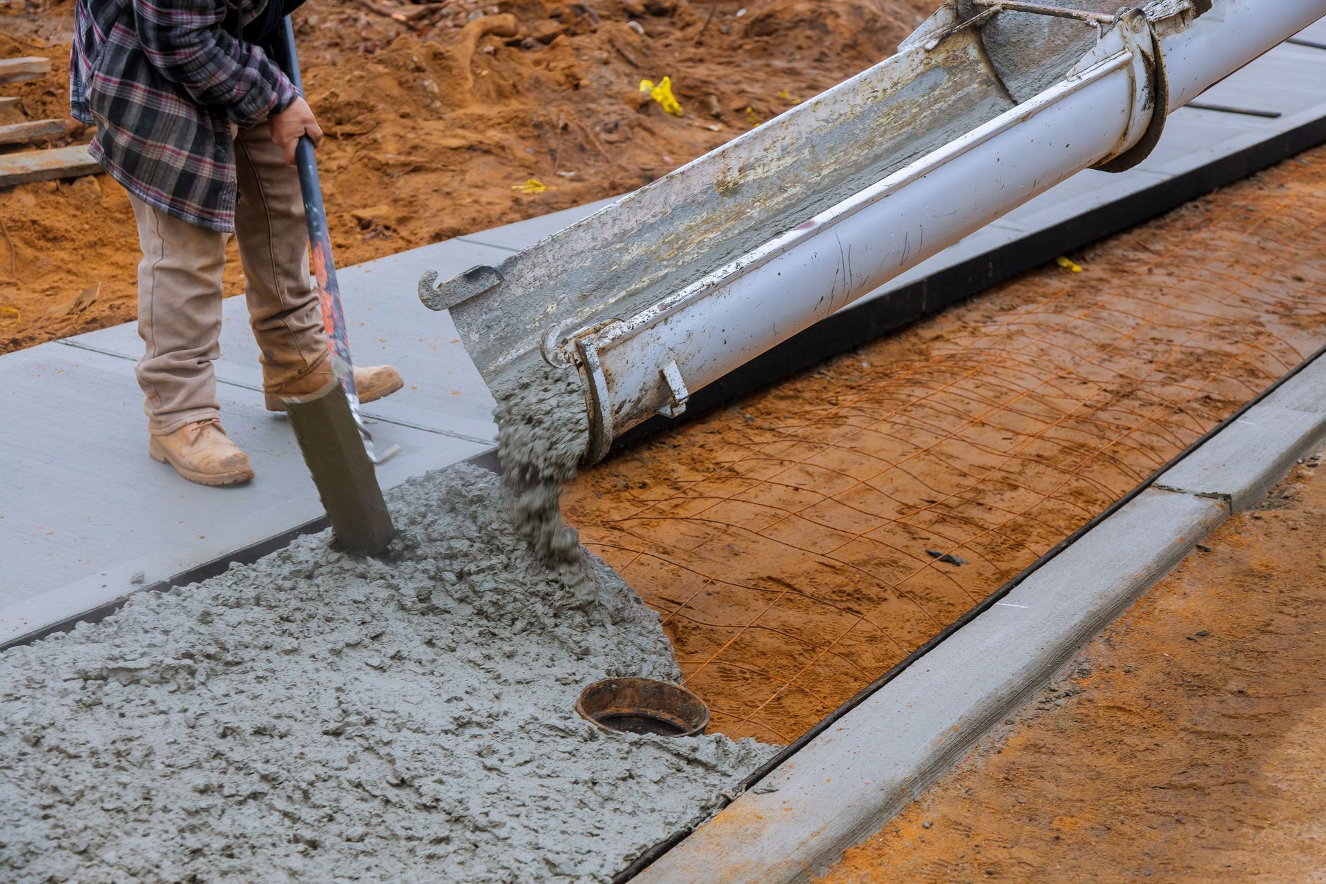 Concrete contractor pouring fresh concrete for sidewalk with chute and leveling tool on site.