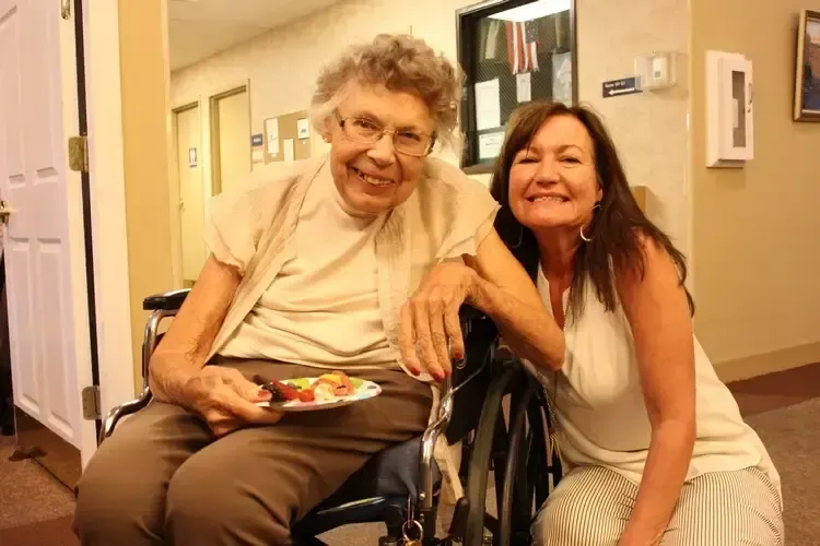 A woman is kneeling next to an elderly woman in a wheelchair holding a plate of food.