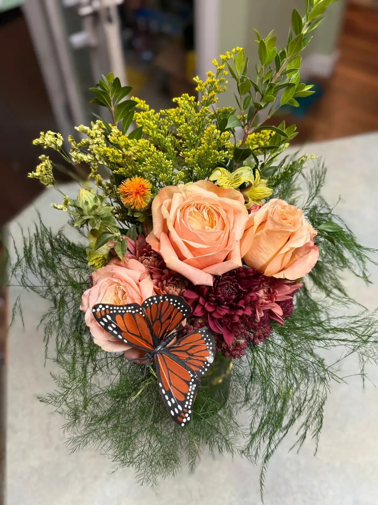 Bouquet of peach roses, maroon dahlias, and greenery, with an orange butterfly.
