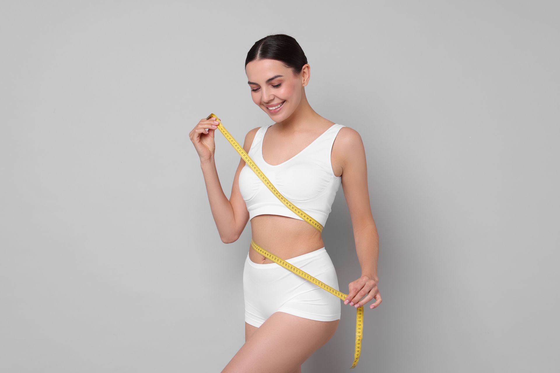 A smiling person in white athletic wear holds a yellow measuring tape wrapped around their torso against a gray background.