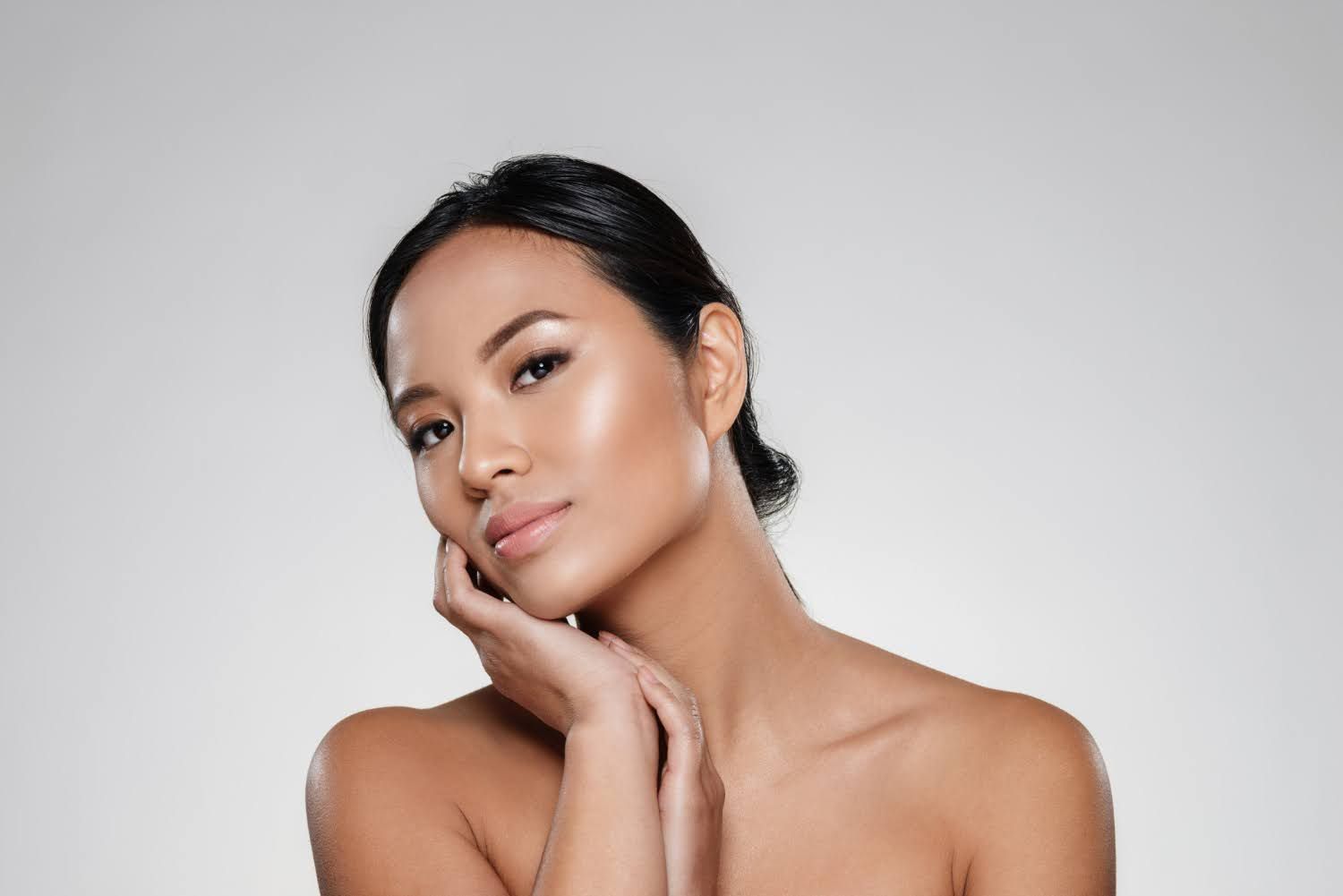 Woman with dark hair and luminous skin, hands near face, looking to the side.