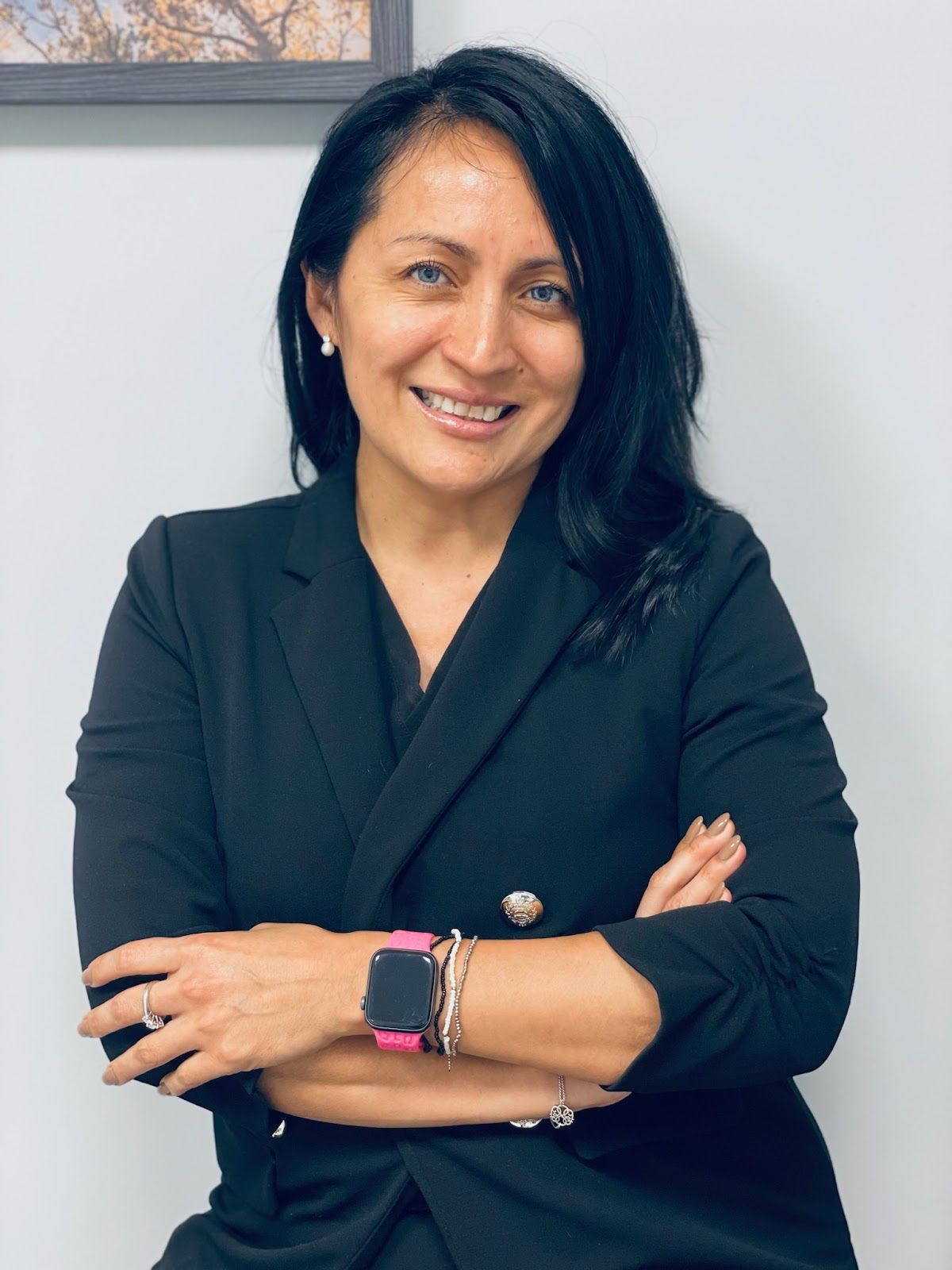 A woman wearing a black jacket and a pink watch is smiling with her arms crossed.